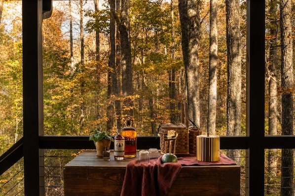 A wooden table with a bottle, glasses, and a plant by a window overlooking a forest. - Sleepy Hollow at The Retreat at Sunset Bluff (Monteagle)
