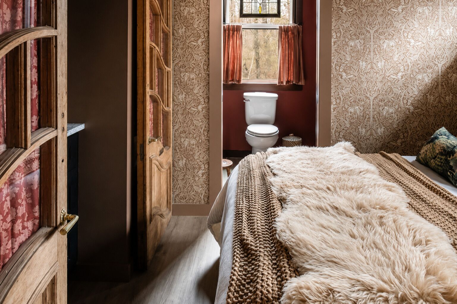 A bedroom with a large bed, a wooden door, a window with curtains, and a toilet.