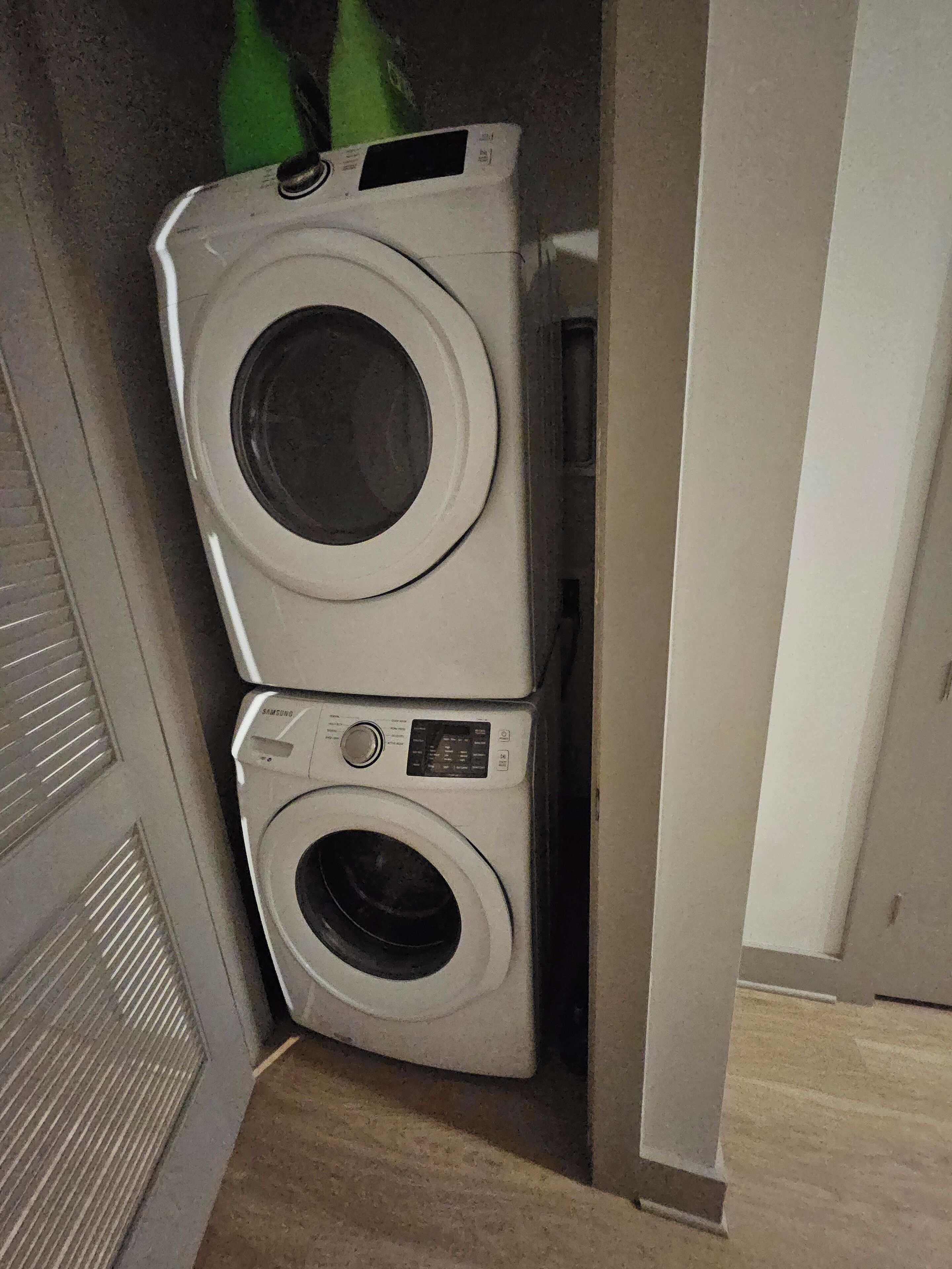 A stacked washer and dryer unit in a laundry room with a vented door and a wooden floor.