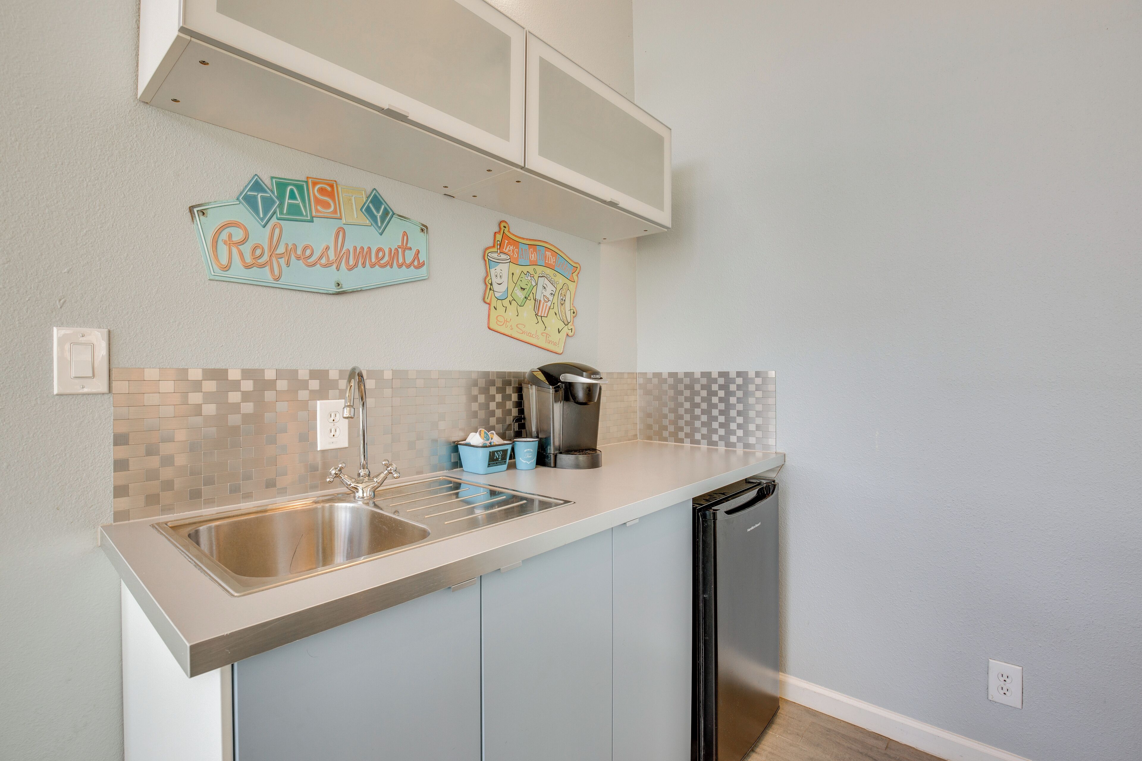 A kitchen with a stainless steel sink, a coffee maker, and a dishwasher. There are two cabinets above the sink and a backsplash with a checkered pattern.