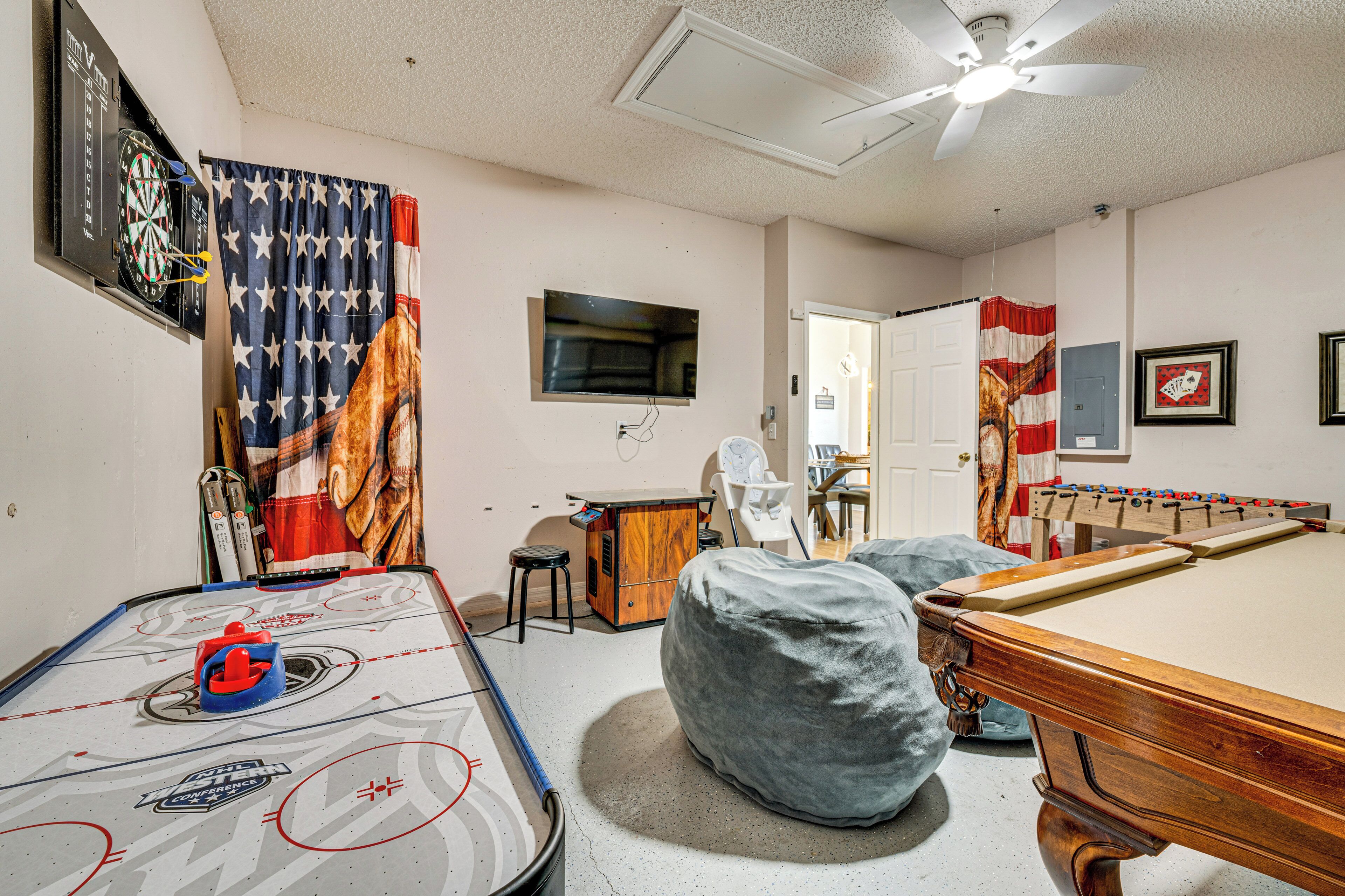 A room with a pool table, a dartboard, a foosball table, and a small bar area.
