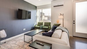 Un salon moderne comprenant un canapé, deux fauteuils, une table basse, un téléviseur à écran plat fixé au mur et une grande fenêtre donnant sur la rue.