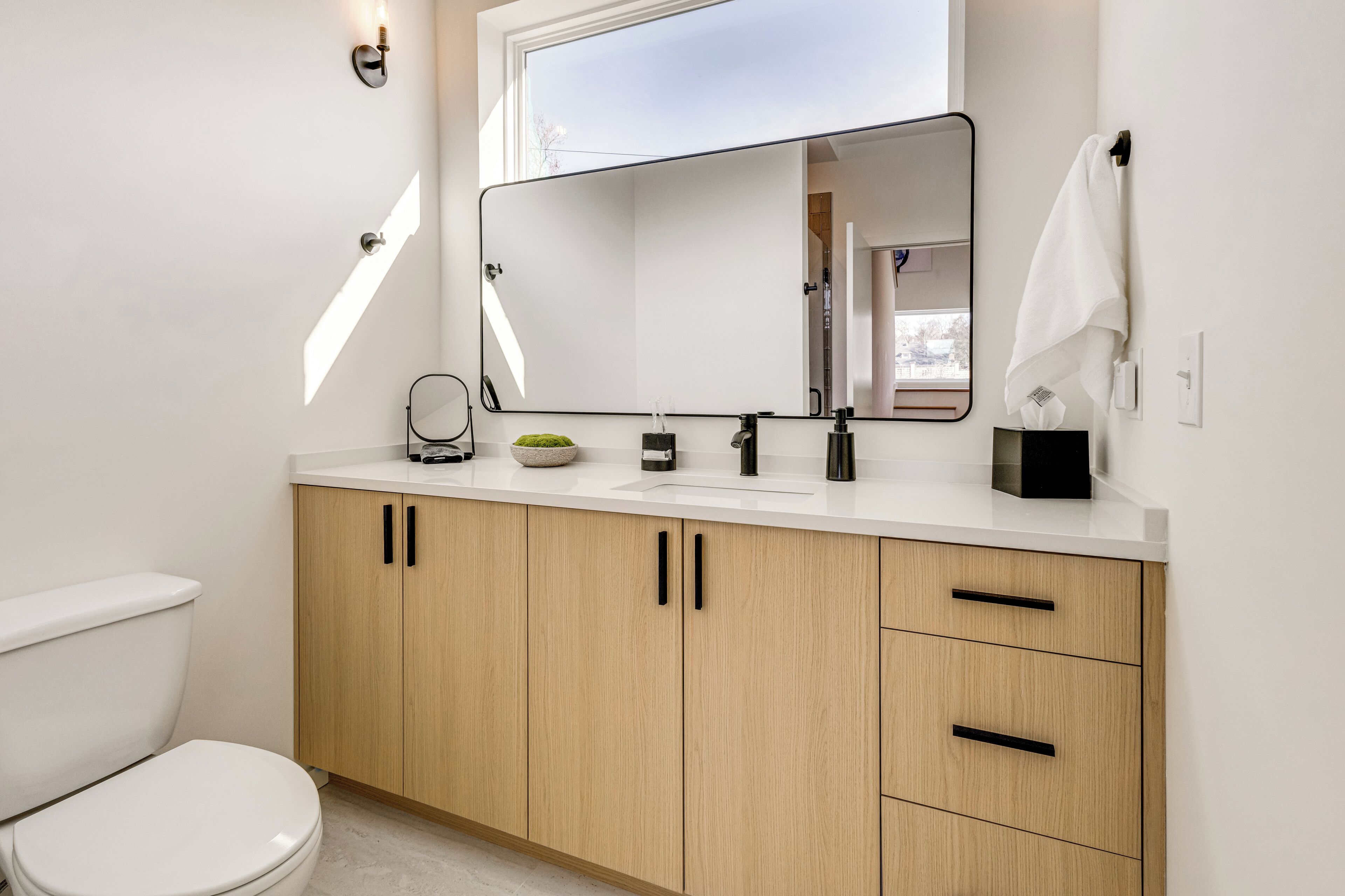 Un baño moderno con inodoro blanco, armarios de madera, un espejo grande y encimera blanca.