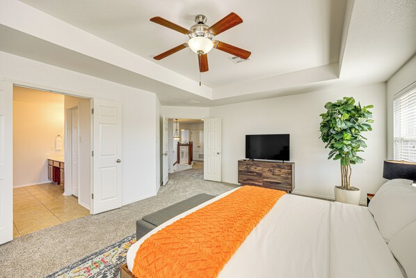 A bedroom with a bed, a ceiling fan, a TV, a plant, and a view of the hallway.