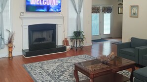 Un salon avec une cheminée, un téléviseur affichant le mot « BIENVENUE », un canapé vert et un tapis à motifs.