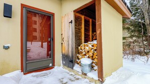 Une entrée enneigée, avec une porte rouge et une porte en bois donnant sur un espace de rangement où se trouvent un seau et une pile de bois de chauffage.