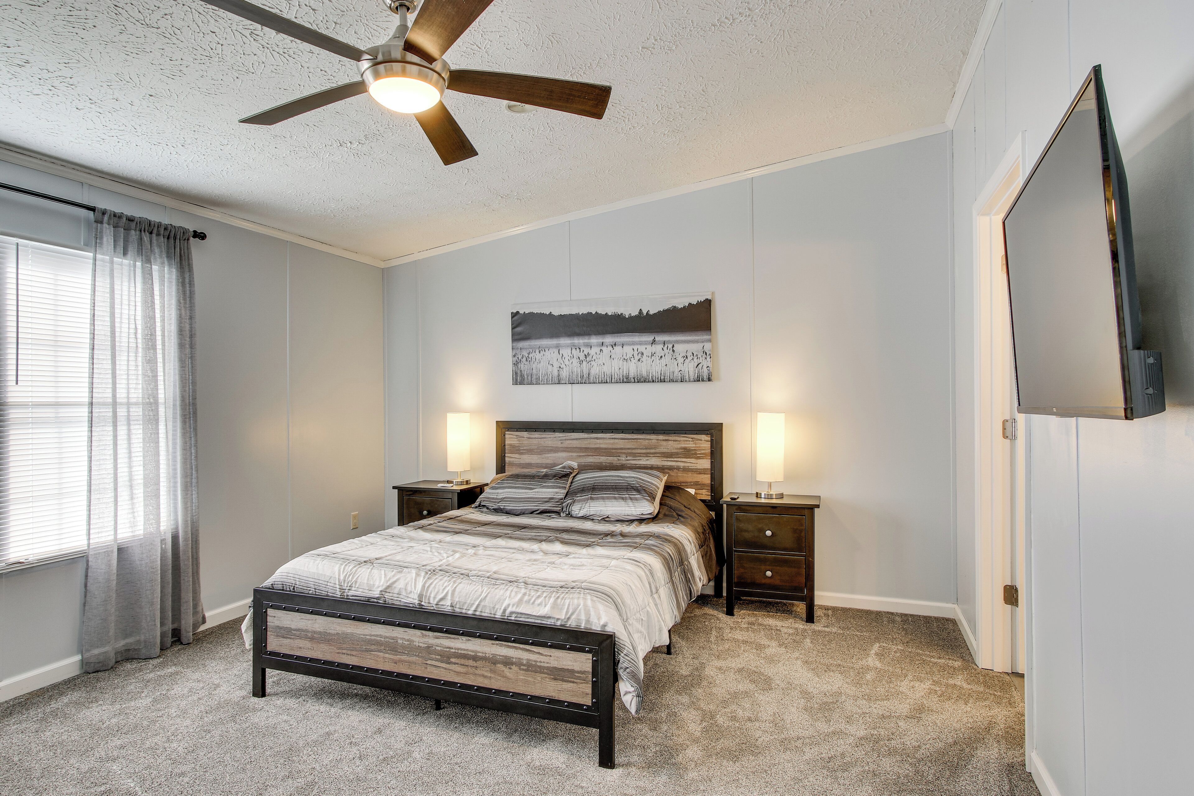 A bedroom with a bed, a ceiling fan, a nightstand, a bedside table, a wall-mounted TV, and a window with curtains.