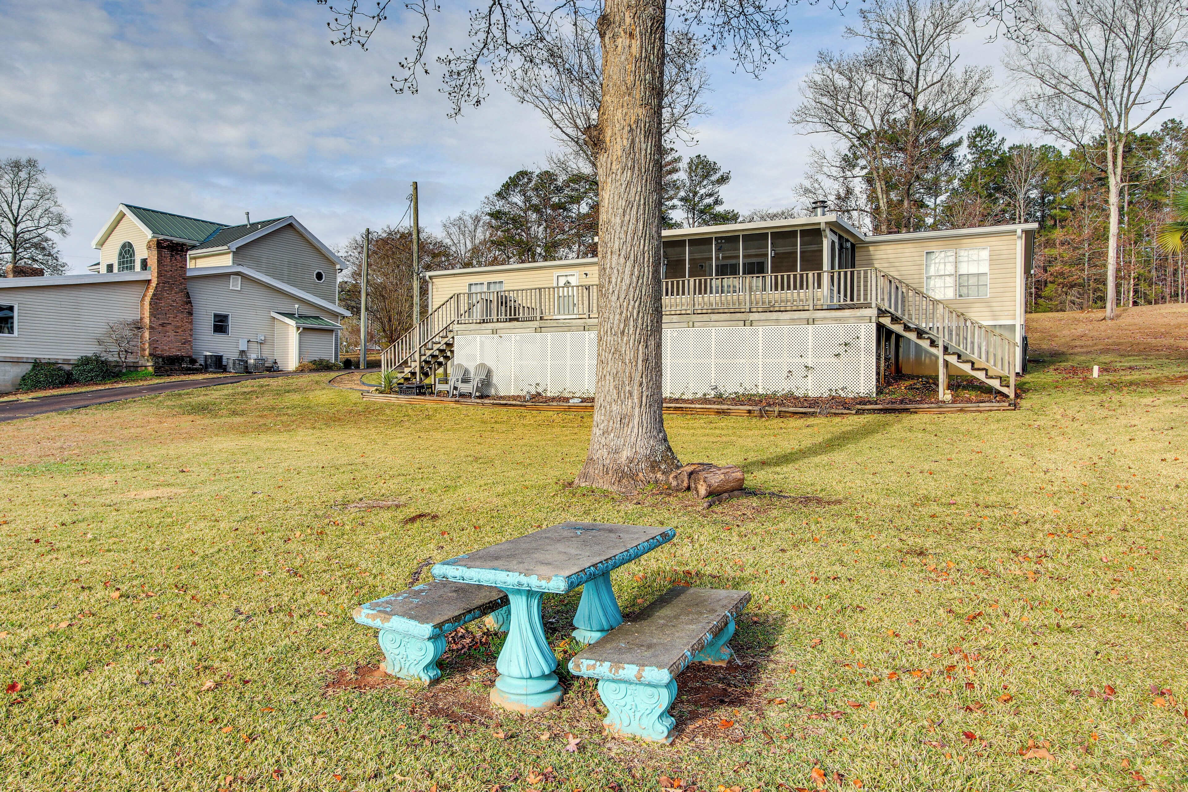 A picnic table with benches in a grassy area, a house with a porch, and a tree in the foreground.