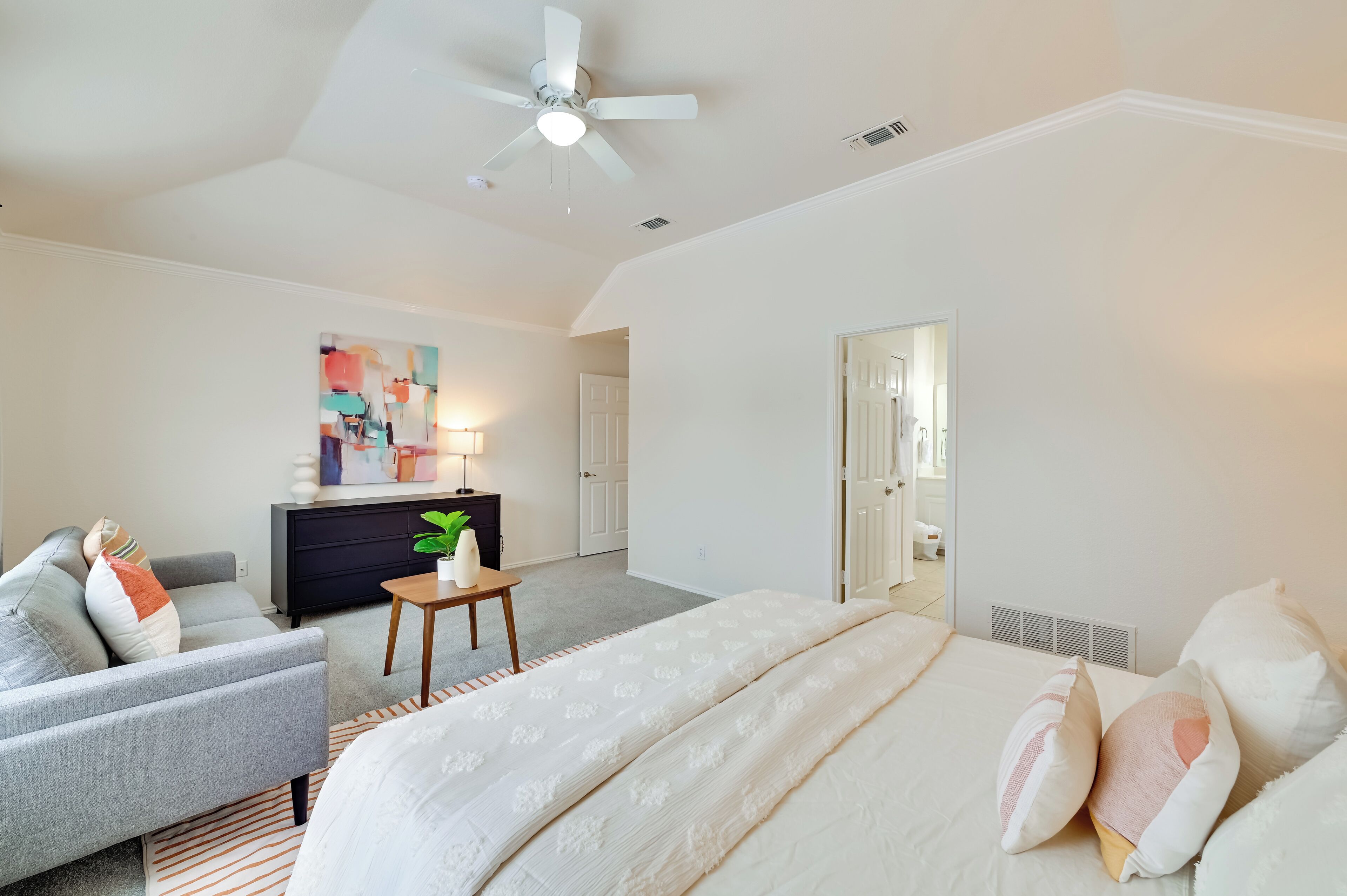 A bedroom with a bed, a gray armchair, a small table with a plant, a dresser, and a ceiling fan.