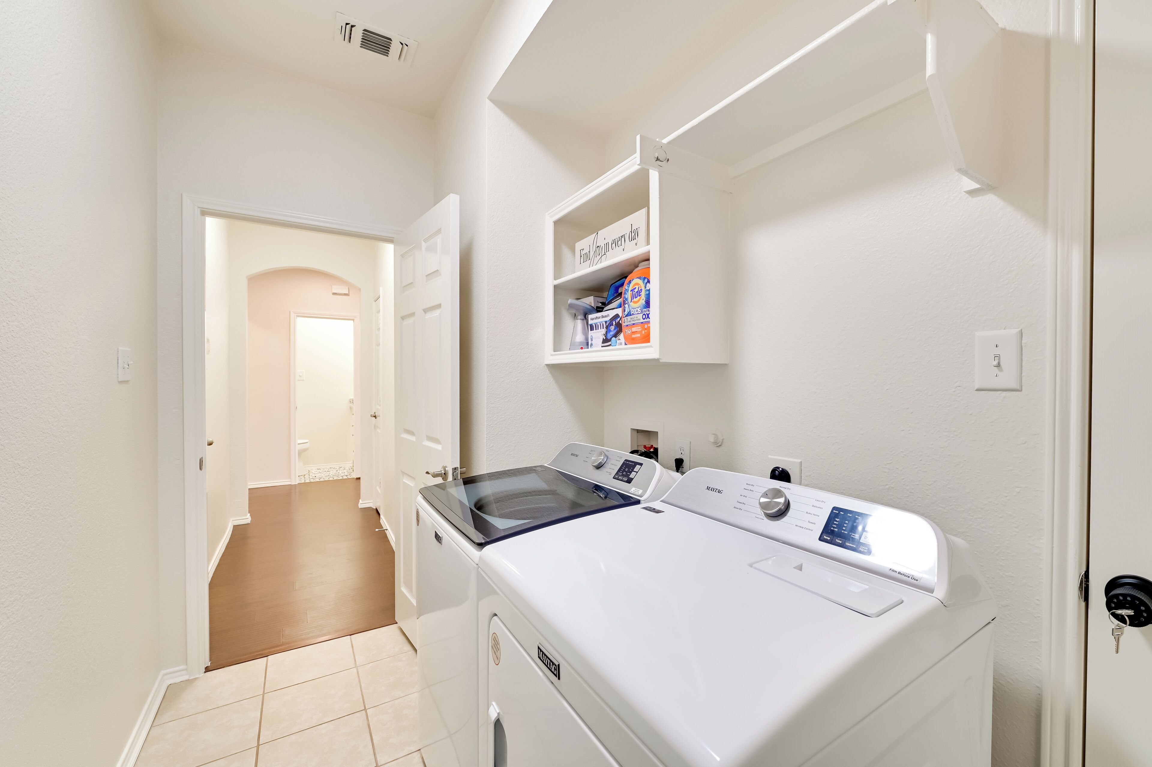 A laundry room with a front-loading washer and dryer, a shelf with laundry supplies, and a door leading to another room.