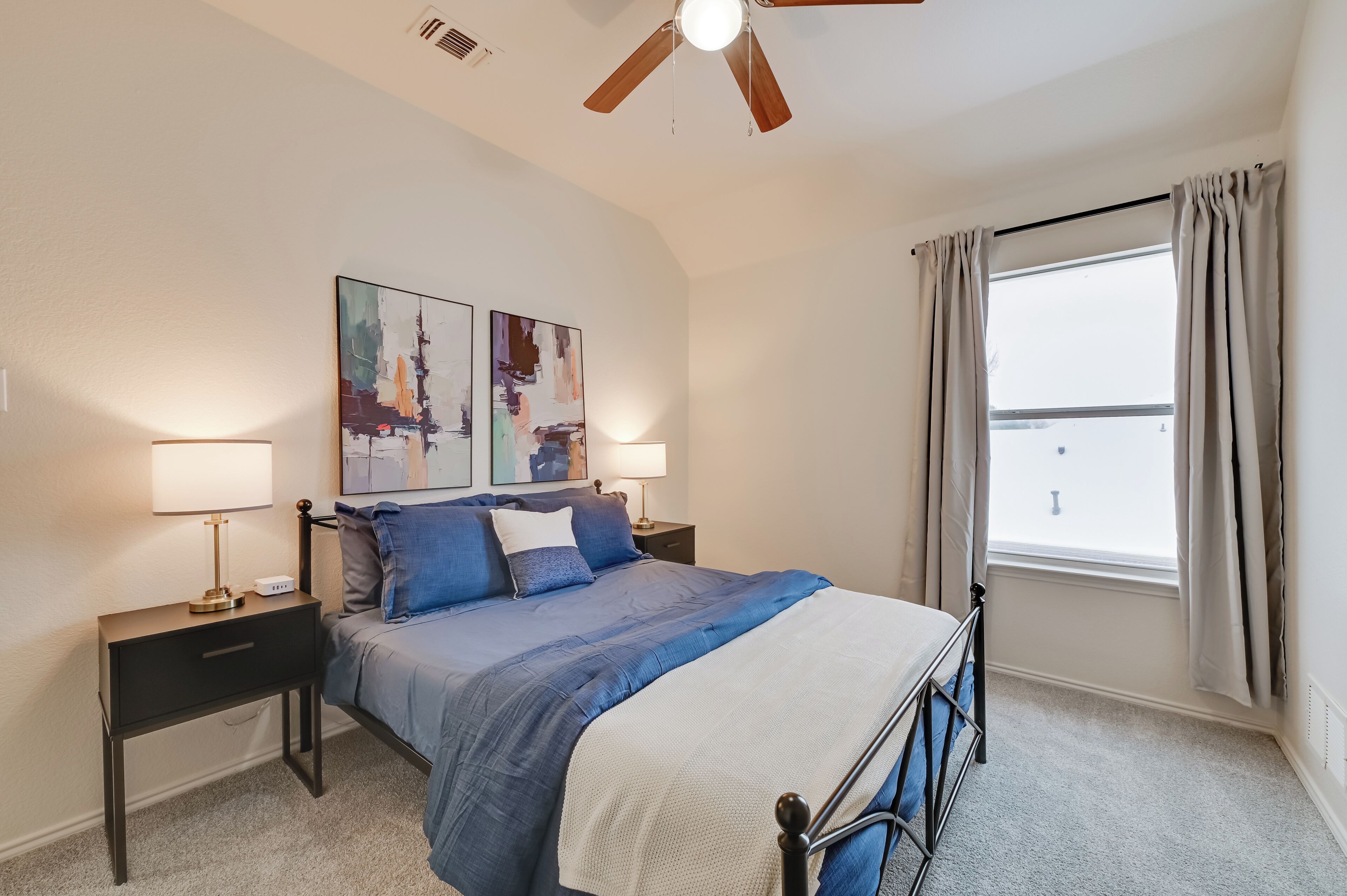 A bedroom with a bed, two bedside tables, a ceiling fan, a window with curtains, and abstract wall art.