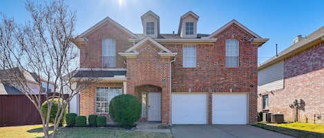 A two-story brick house with a front garage, a small front yard with a tree, and a clear blue sky.