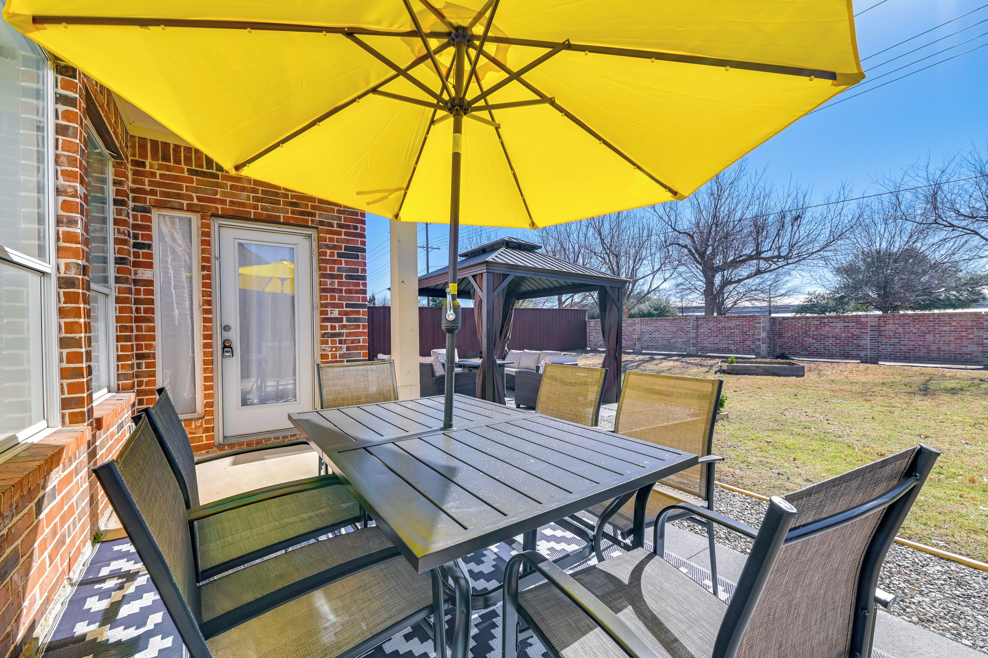 Een terras met een tafel en stoelen, een gele parasol, en een stenen huis op de achtergrond.