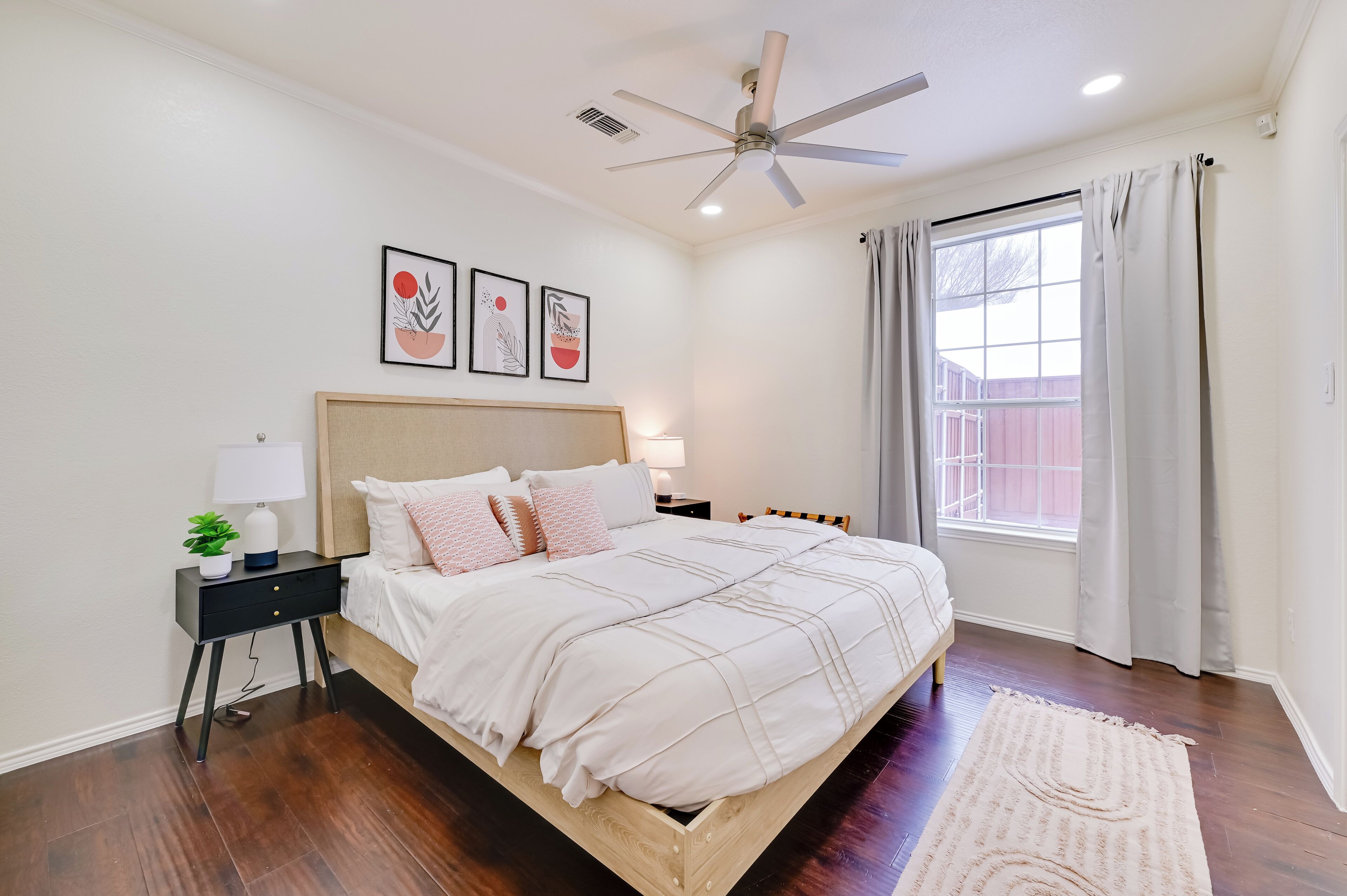 A bedroom with a bed, bedside table, lamp, ceiling fan, and artwork on the wall.