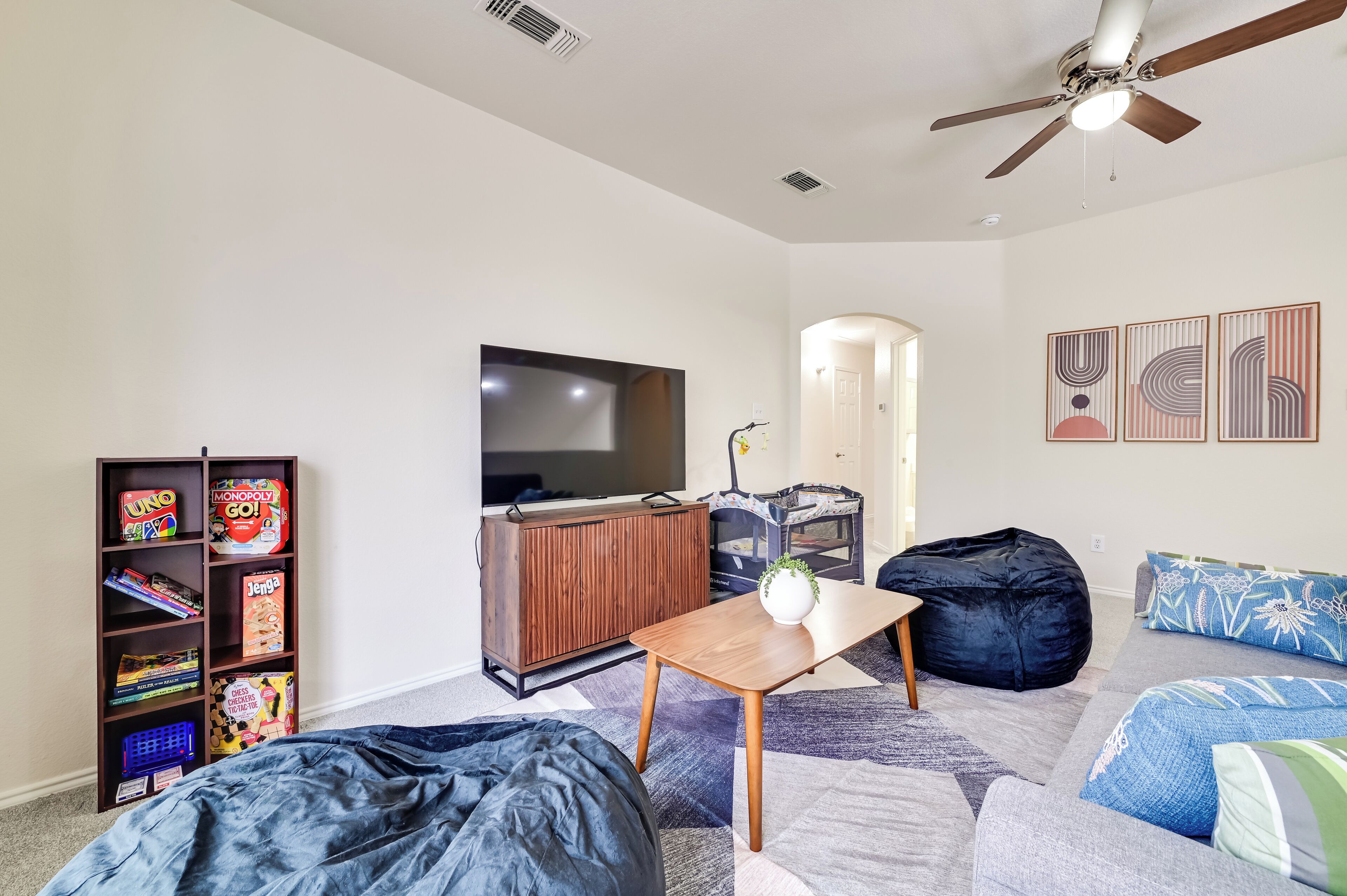 A living room with a flat-screen TV, a bookshelf with board games, a wooden coffee table, a bean bag, and a gray sofa.
