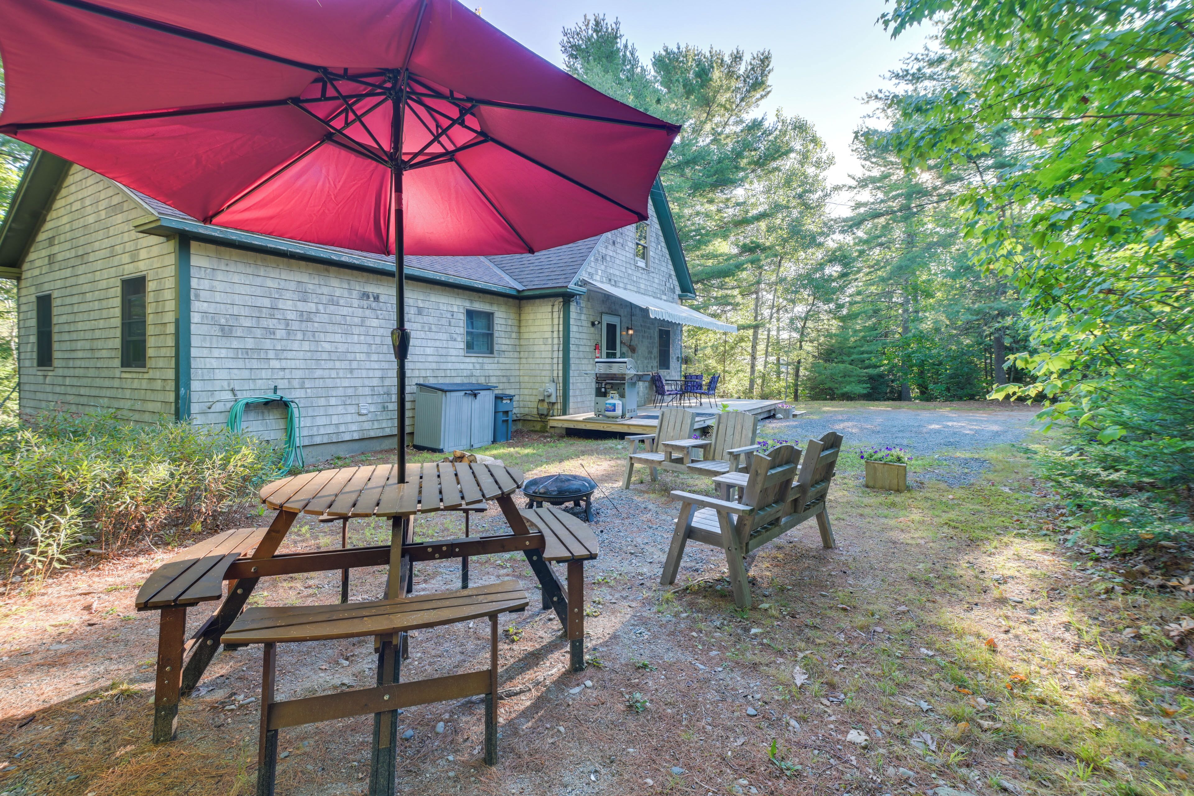 A wooden deck with picnic tables and chairs, a red umbrella, and a house in the background.