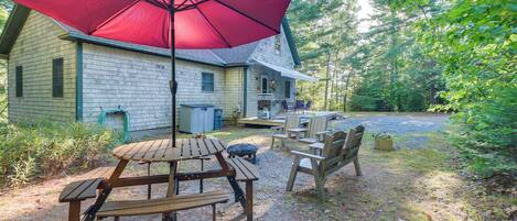 A wooden deck with picnic tables and chairs, a red umbrella, and a house in the background.