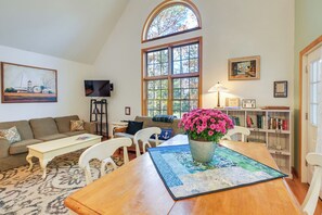A cozy living room with a high arched window, a wooden dining table with a blue placemat, a white sofa, and a bookshelf filled with books. - 17 Mi to Acadia: Cottage Near Dtwn Ellsworth! (Ellsworth)