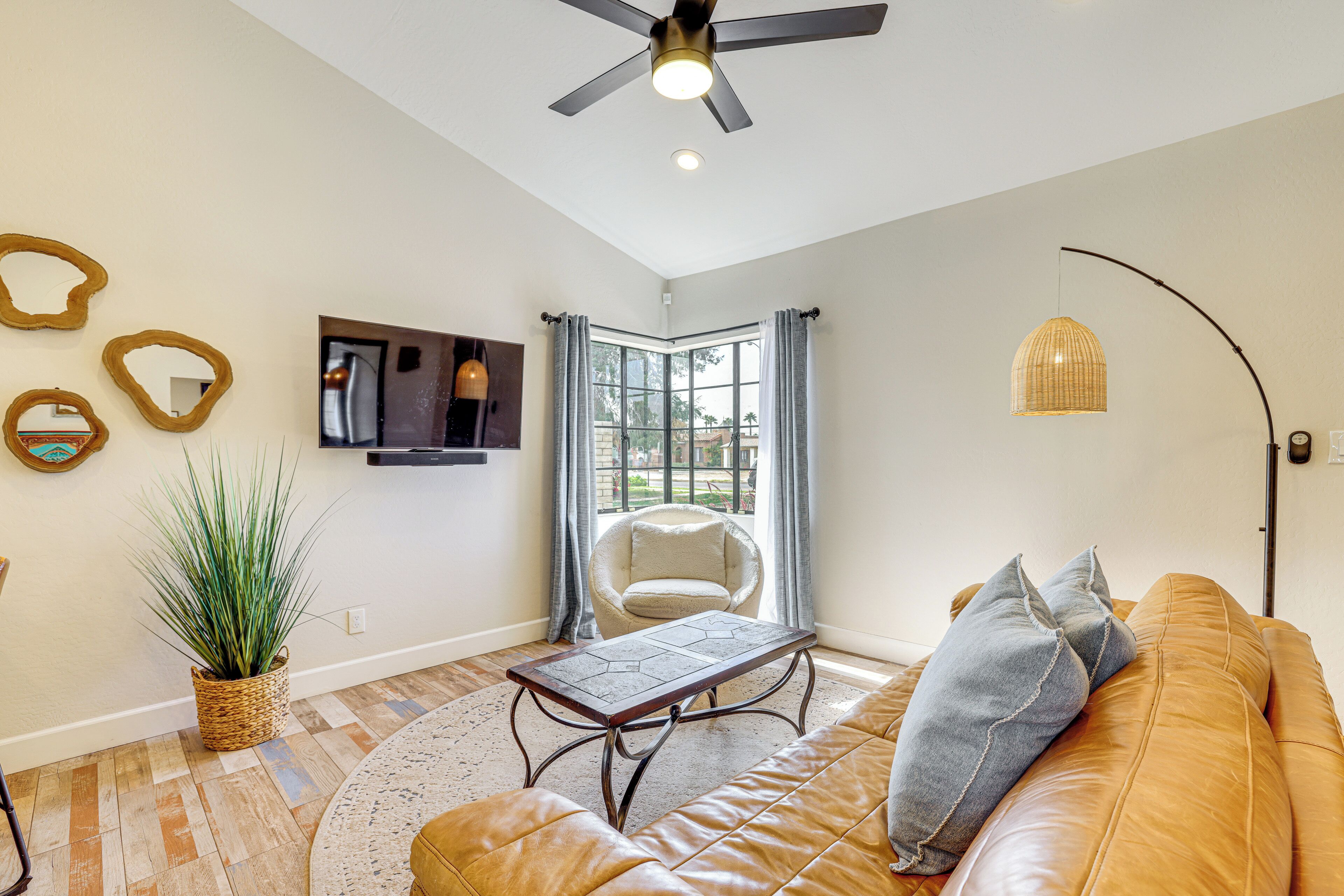 A living room with a leather sofa, a coffee table, a TV mounted on the wall, a ceiling fan, and a large window with curtains.