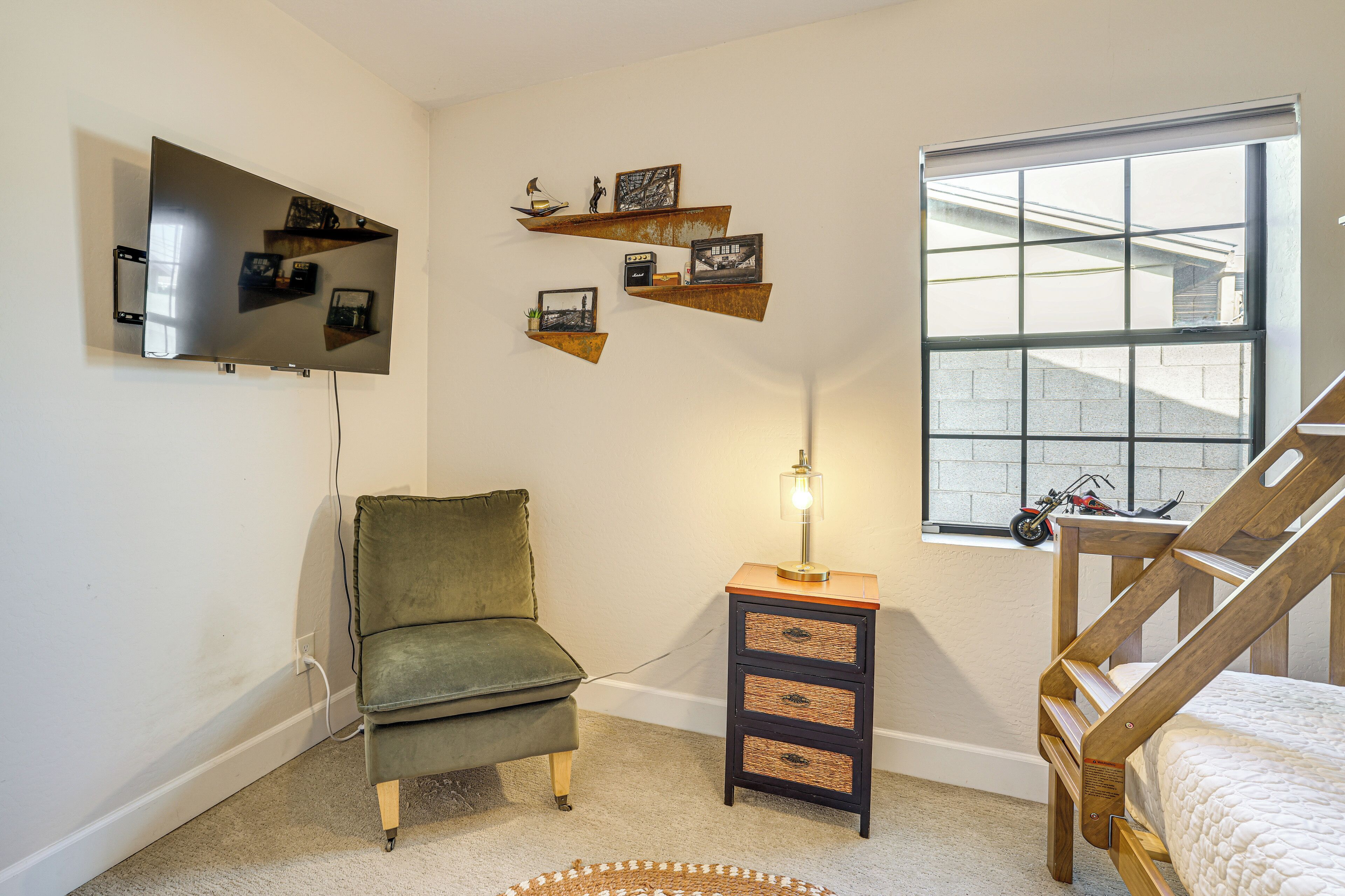 A living room with a green armchair, a wooden dresser, a TV mounted on the wall, and a window with a view of a building outside.