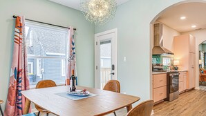 A dining area with a wooden table and chairs, a kitchen in the background, and a view of the outside through a window.