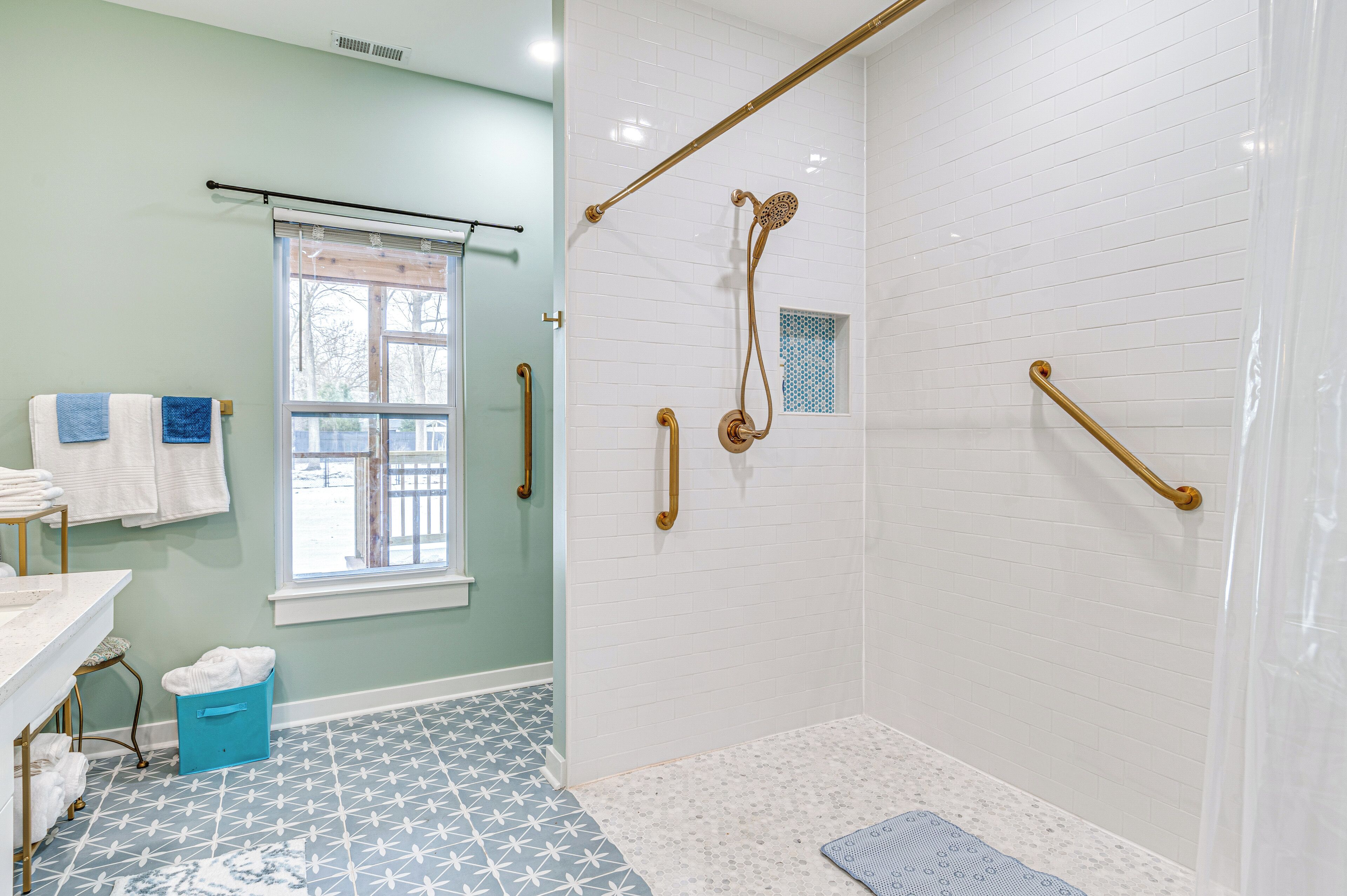 A bathroom with a shower, grab bars, a window, and a blue trash bin.