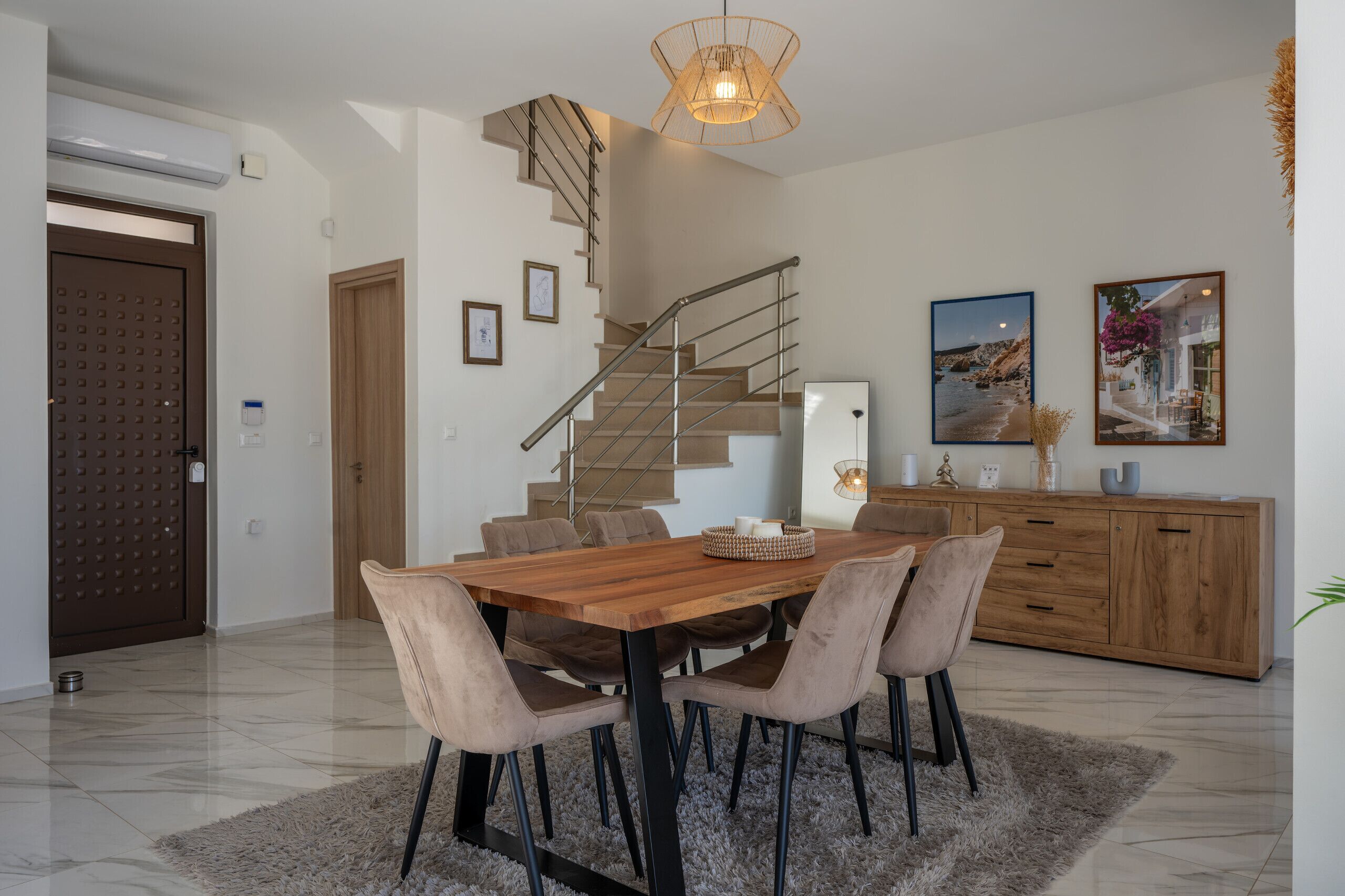 A modern dining area with a wooden table, cushioned chairs, a staircase, and a view of the outdoors.
