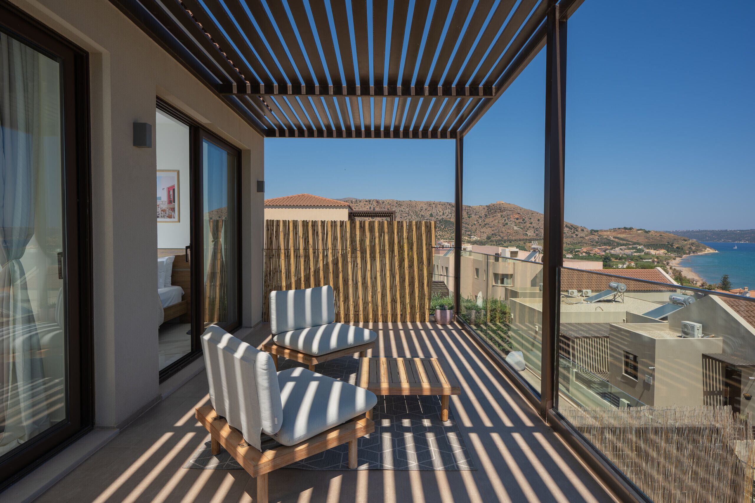 A balcony with two striped chairs, a small table, and a view of the sea and hills.