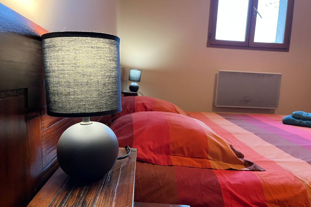 A bed with a colorful quilt, a bedside lamp, a wooden nightstand, a small table with a lamp, and a window with blinds.