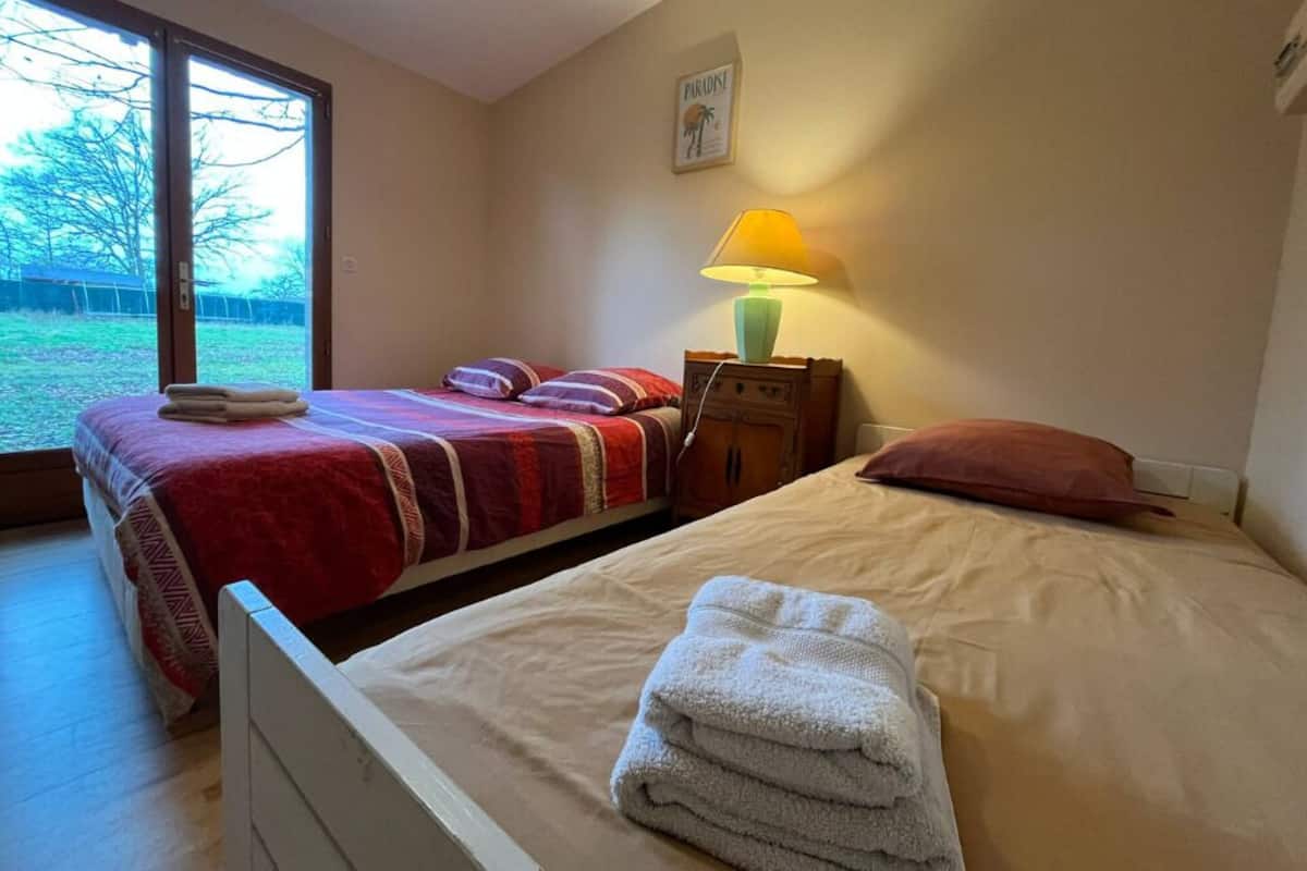 Une chambre avec deux lits, une fenêtre donnant sur des arbres, une lampe et un tableau encadré au mur.