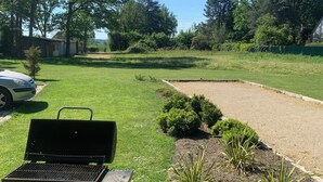 A grassy outdoor area with a barbecue grill, a paved path, and a small building in the background. - Maison du verger idéal famille/ami & séjour pro (Rives-du-Loir-en-Anjou)