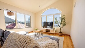 Ein Wohnzimmer mit großem Fenster, einem Flachbildfernseher, einer Couch, einem Couchtisch und einer Topfpflanze.