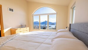 Ein Schlafzimmer mit einem großen Bett, einer Holzkommode, einem Fenster mit Blick auf Berge und Wasser sowie einem kleinen Tisch mit einem Korb.