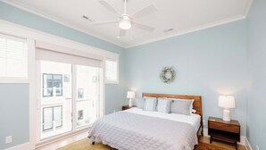Une chambre à coucher comprenant un lit, des tables de chevet, un ventilateur de plafond, une fenêtre avec des stores et une porte.
