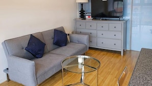 Un salon avec un canapé gris, deux coussins bleus à motifs, une table basse en verre, un téléviseur posé sur un meuble gris et un plancher en bois.