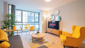 Ein modernes Wohnzimmer mit Sofa, Sessel und Couchtisch, ein Essbereich mit Stühlen und Tisch sowie ein an der Wand befestigter Fernseher.