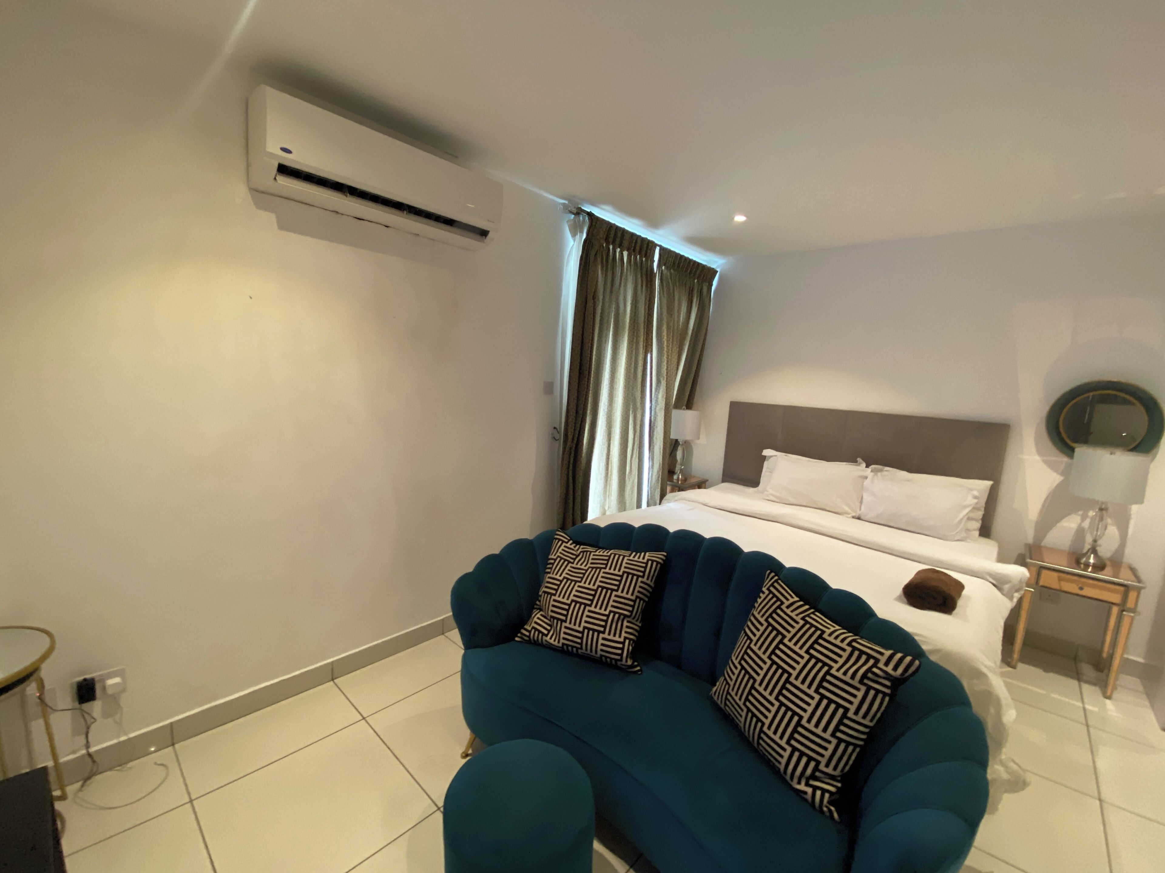 A modern hotel room with a large bed, a sitting area with a blue sofa, and an air conditioning unit on the wall.