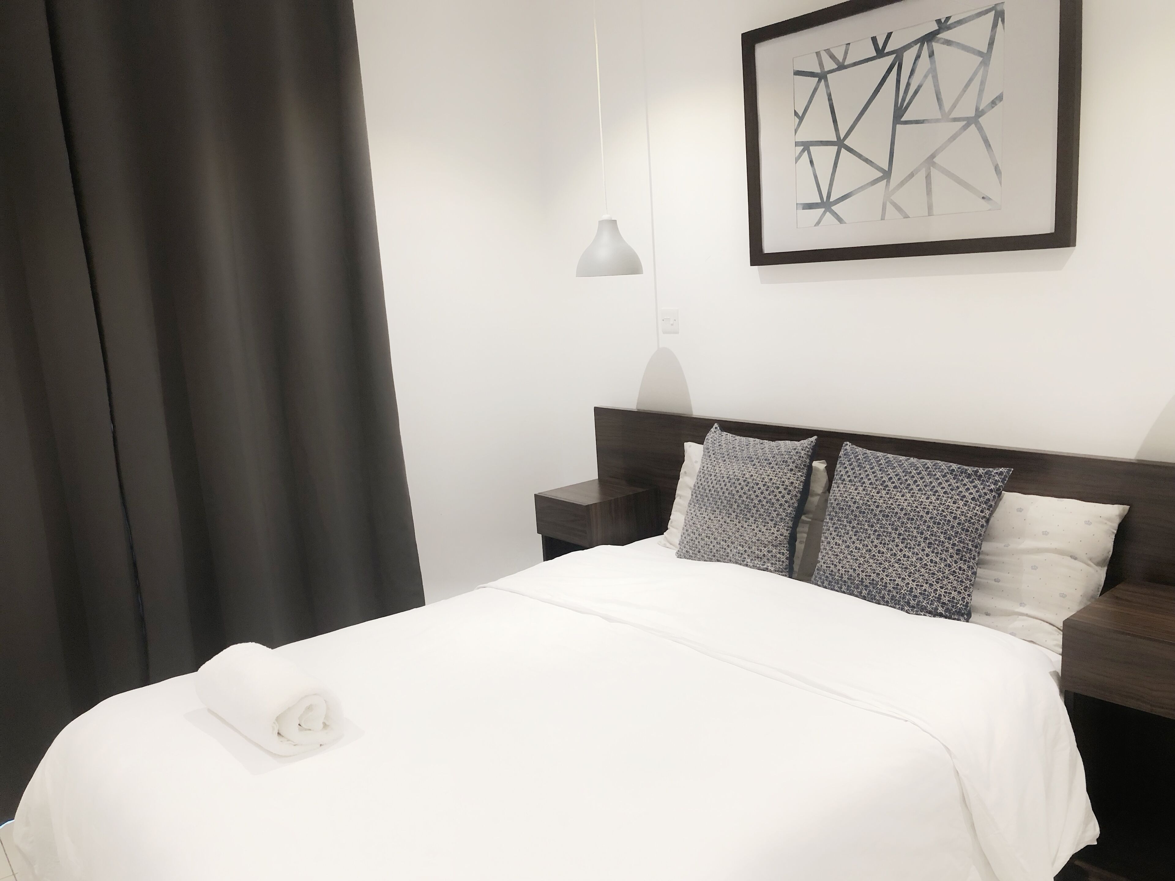 A neatly made bed with white bedding and two patterned pillows, a dark wooden headboard, a small bedside table, and a modern abstract painting on the wall.