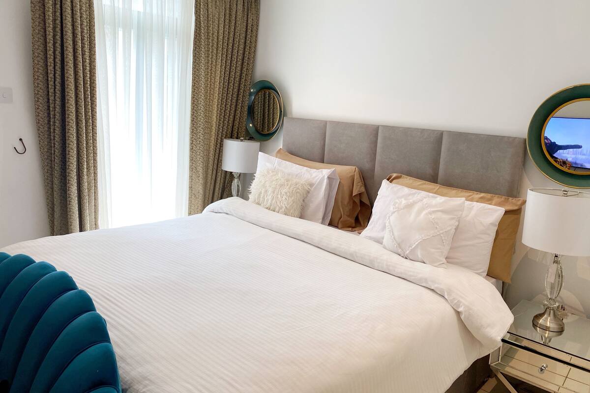 A neatly made bed with white linens, a grey upholstered headboard, and a bedside table with a lamp.