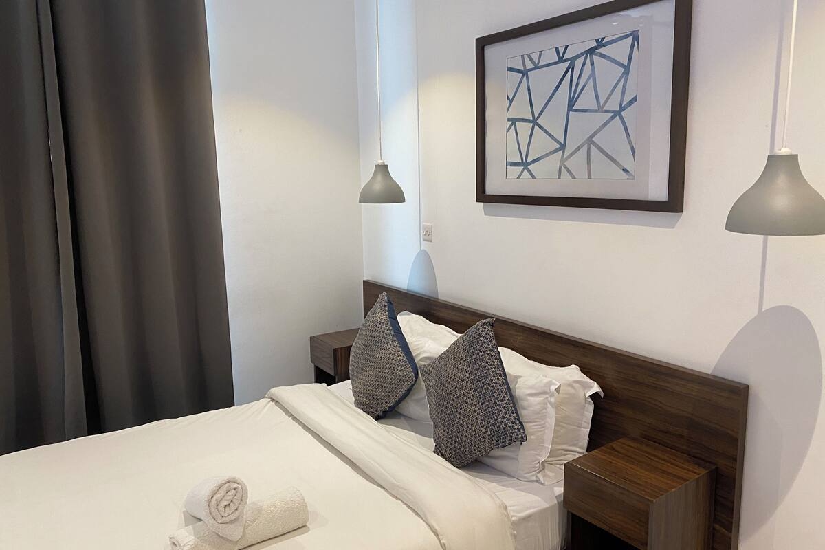 A neatly made bed with a rolled towel, two pillows, a framed abstract artwork, and two bedside lamps.