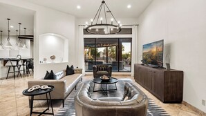 A modern living room with a large sofa, a coffee table, and a TV on a wooden cabinet. There is a dining area with a table and chairs, and a kitchen visible through an open door. - Private Movie Theater, Luxury Pool, Hot Tub (Scottsdale)