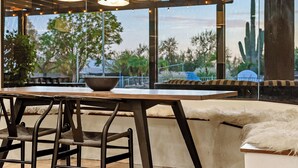 An outdoor seating area with a wooden table and chairs, a bowl on the table, and a view of trees and a cactus through large windows. - Private Movie Theater, Luxury Pool, Hot Tub (Scottsdale)