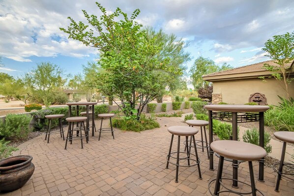 An outdoor patio with multiple bar stools and tables, surrounded by greenery and a clear sky.