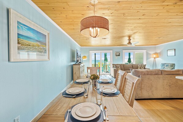 Un coin repas avec une table en bois dressée pour six personnes, un lustre et une vue sur l’extérieur.