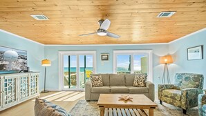 A living room with a wooden ceiling, a sofa, a coffee table, and a view of the beach through large windows.