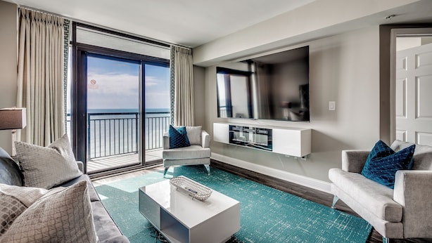 A modern living room with a sofa, coffee table, and TV. A balcony with curtains is visible. - 🌊Newly Renovated Oceanfront | Awesome Amenities | Caribbean Resort 1405🌊 (Myrtle Beach)