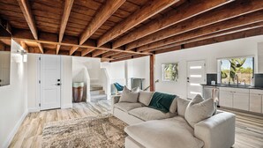 Un vaste espace de vie doté d’un haut plafond en bois, d’un canapé beige, d’un tapis moelleux et d’un coin cuisine avec une fenêtre.