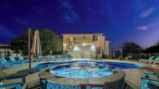 Un espace piscine doté de chaises longues et d’un jacuzzi, le tout entouré d’un bâtiment illuminé de guirlandes lumineuses.
