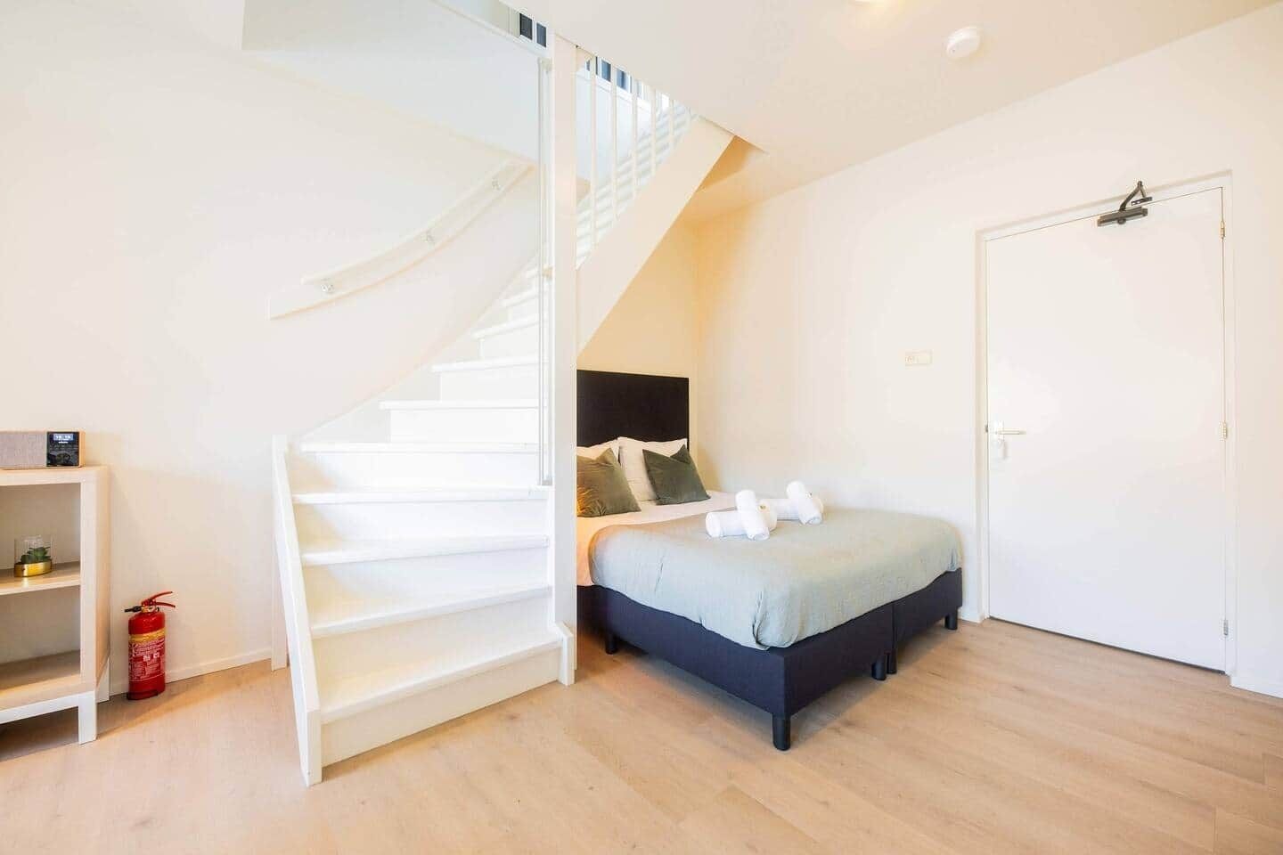 Ein Schlafzimmer mit einem Bett, einer weißen Tür, einer weißen Treppe und einem kleinen Regal mit einem Feuerlöscher.