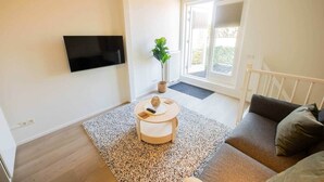 Una sala de estar moderna con un televisor de pantalla plana montado en la pared, una mesa de centro redonda de madera, un sofá gris y un ventanal con vistas a zonas verdes.